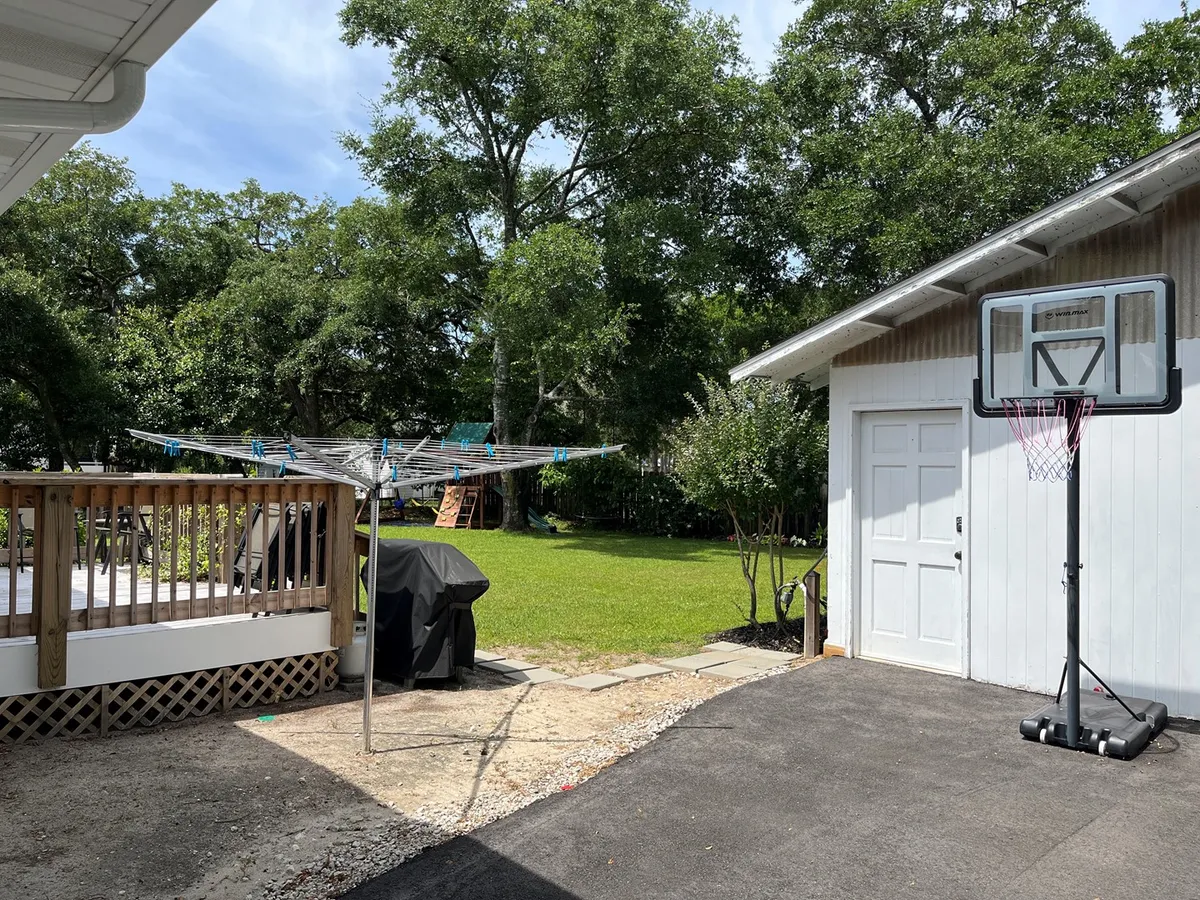 A relaxed exterior scene featuring a basketball hoop, an inviting green lawn, a garage, and a spacious deck area in the background. The deck seems ideal for gathering with friends or catching a sunset. The atmosphere is bright and welcoming, with the space clearly designed for outdoor leisure and socializing.