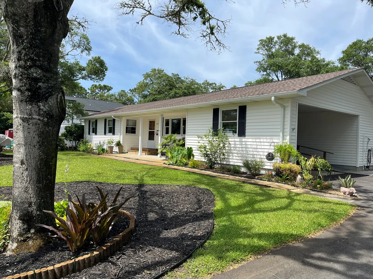 This image depicts the front porch of the house, surrounded by a well-maintained lawn and colorful flower beds. The porch is inviting and evokes a sense of relaxation and community, with plenty of space for guests to gather and enjoy the outdoors. The setting captures the home as a comfortable and welcoming base for summer festivities.