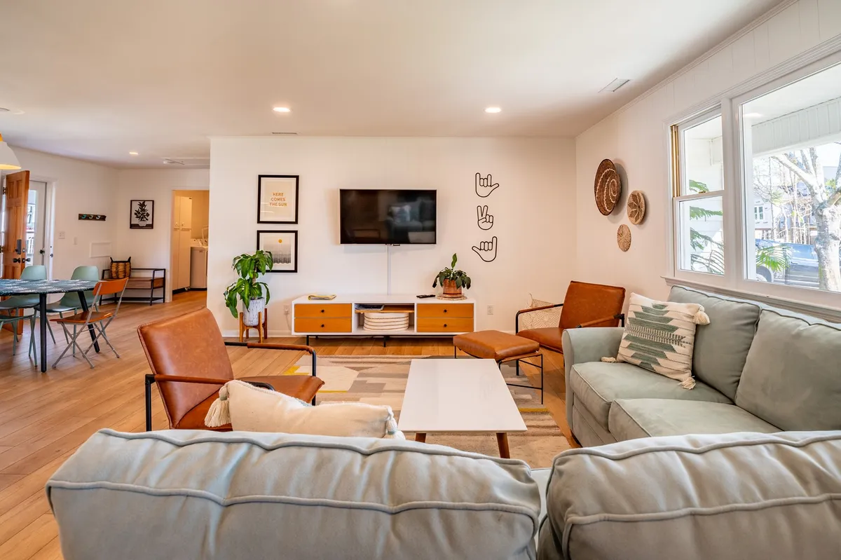 A cozy, well-lit living area featuring a spacious sectional sofa, a coffee table, decorative wall art, a lush green plant, and a smart TV. The natural light streaming in and the open arrangement make it both inviting and ideal for social gatherings.