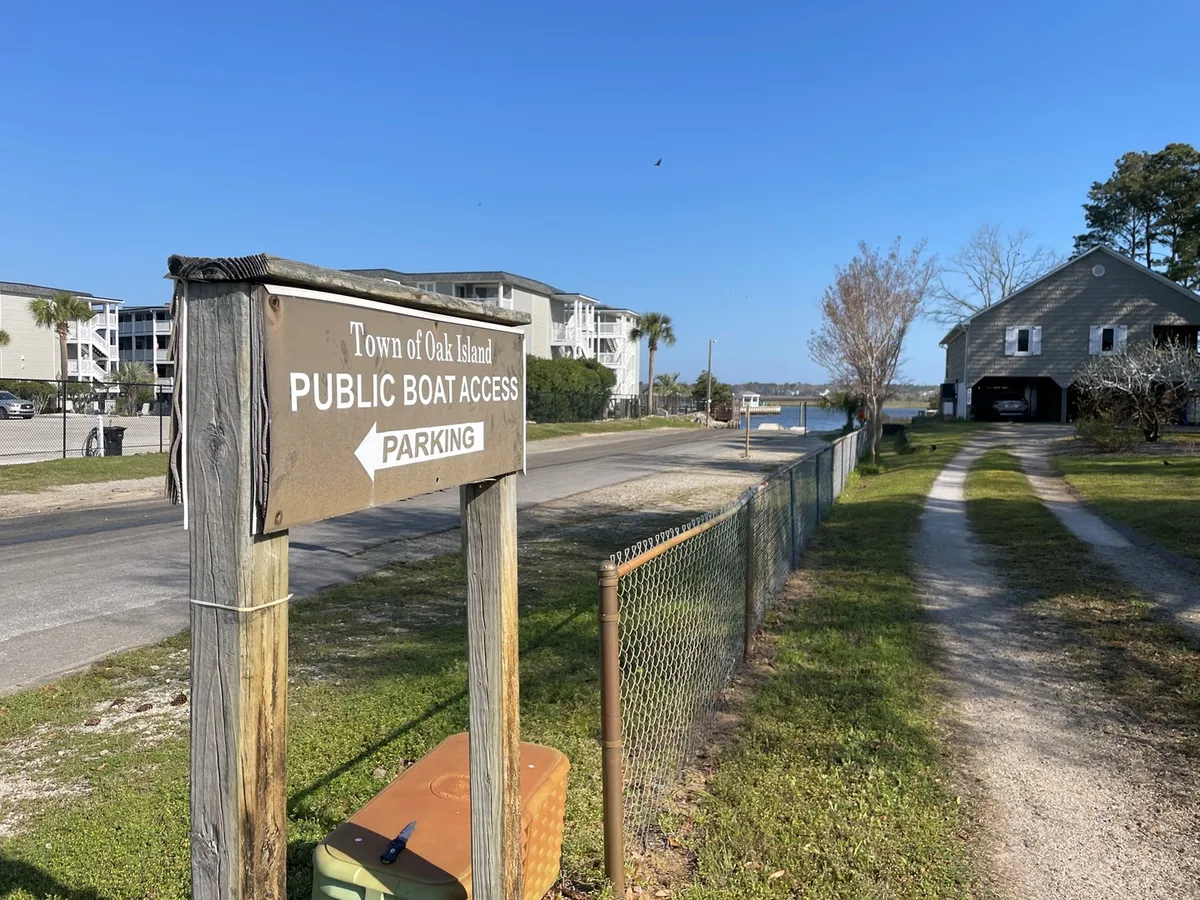 This exterior shot features a public boat access sign, a parking area, and nearby residential buildings. It emphasizes the proximity to local boating, fishing, and water activities, as well as the practical convenience of parking for multiple vehicles and trailers.