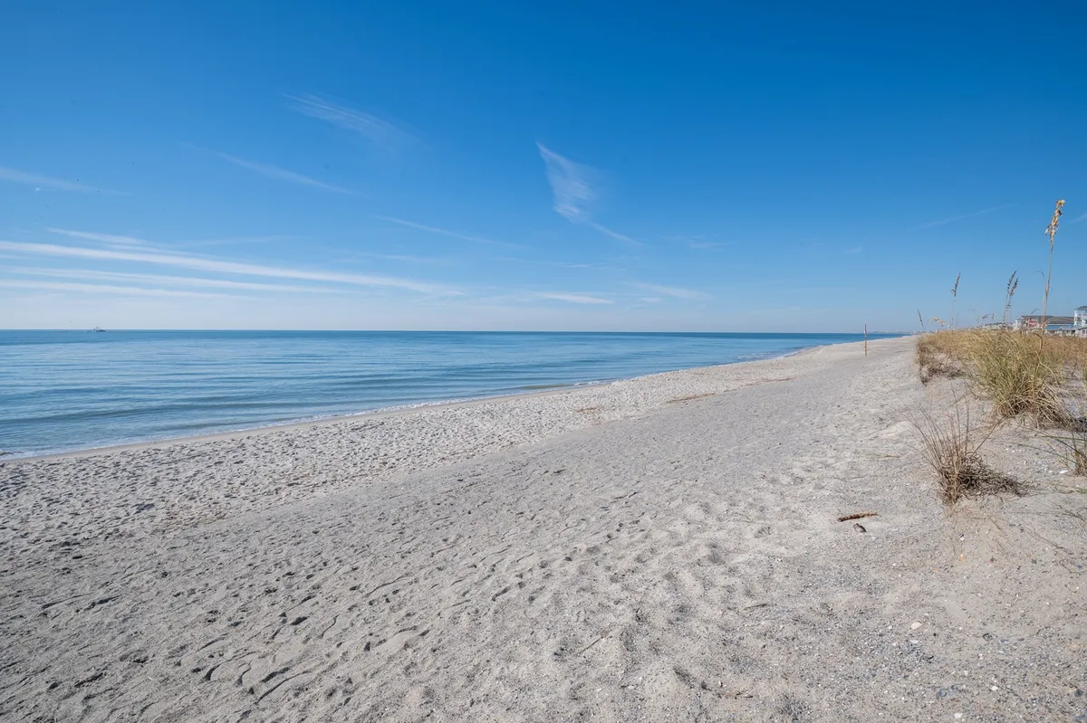 A picturesque view of a wide sand beach with calm sea and a blue sky overhead. The open shoreline is perfect for sunbathing, strolling, and building sandcastles, capturing the tranquil essence of a classic beach day.