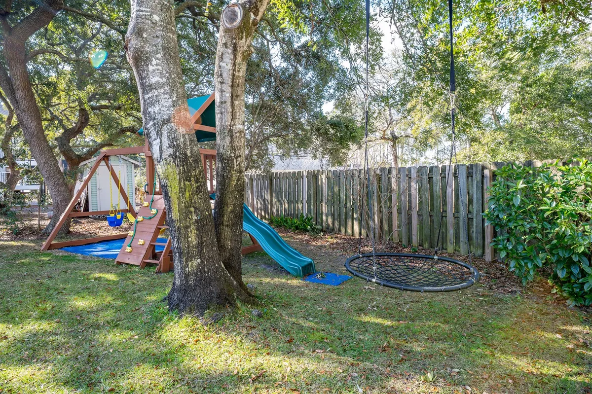 A vibrant outdoor playground surrounded by lush trees, featuring swings, slides, a hammock swing, and picnic areas. Children are seen playing and families gather under the shade, creating an atmosphere of fun and togetherness.