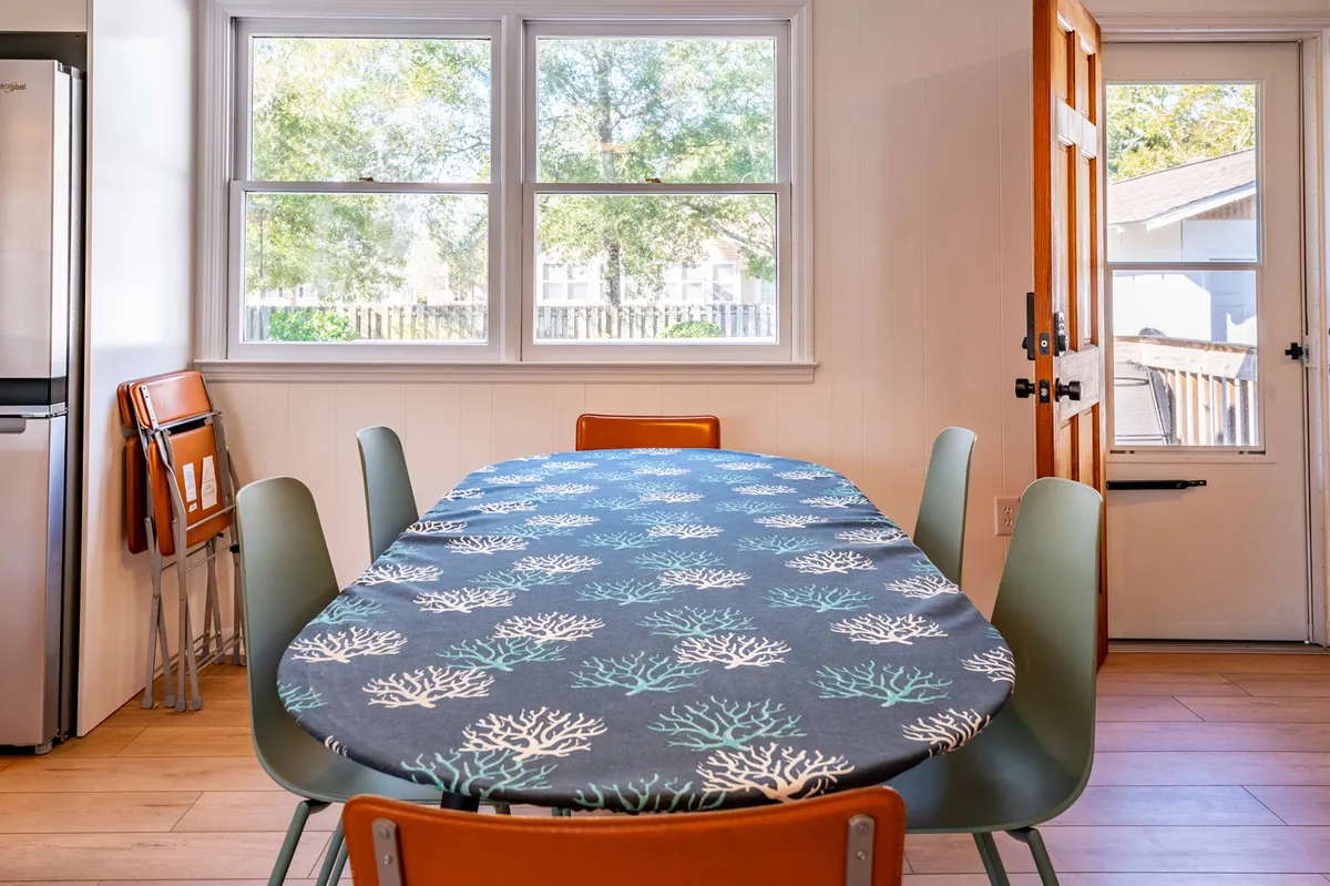 A dining area with a large oval table covered in a coral-patterned tablecloth, surrounded by chairs and bathed in natural light from expansive windows. The scene conveys warmth and togetherness, with a door leading to the outdoors in the background.