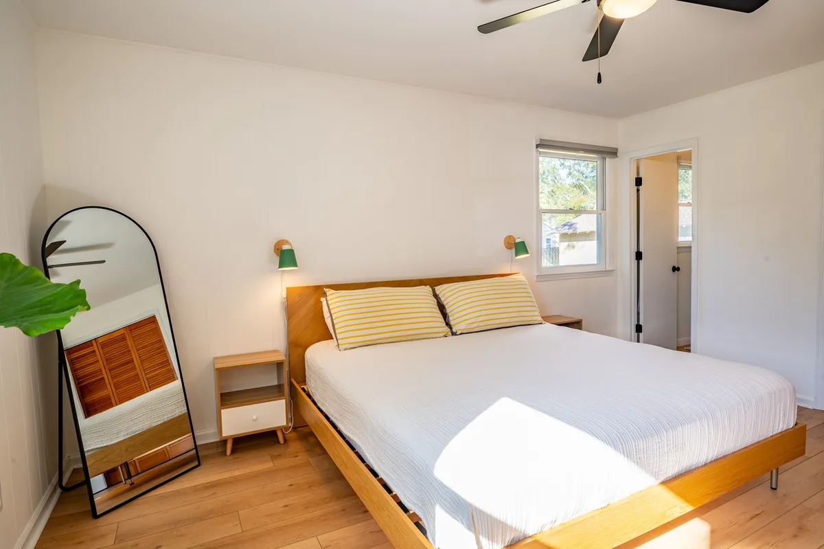 A serene bedroom with a plush bed, nightstand, mirror, and a large window streaming in natural sunlight. The calming palette and uncluttered decor create a relaxing atmosphere, perfect for unwinding after a day at the beach.