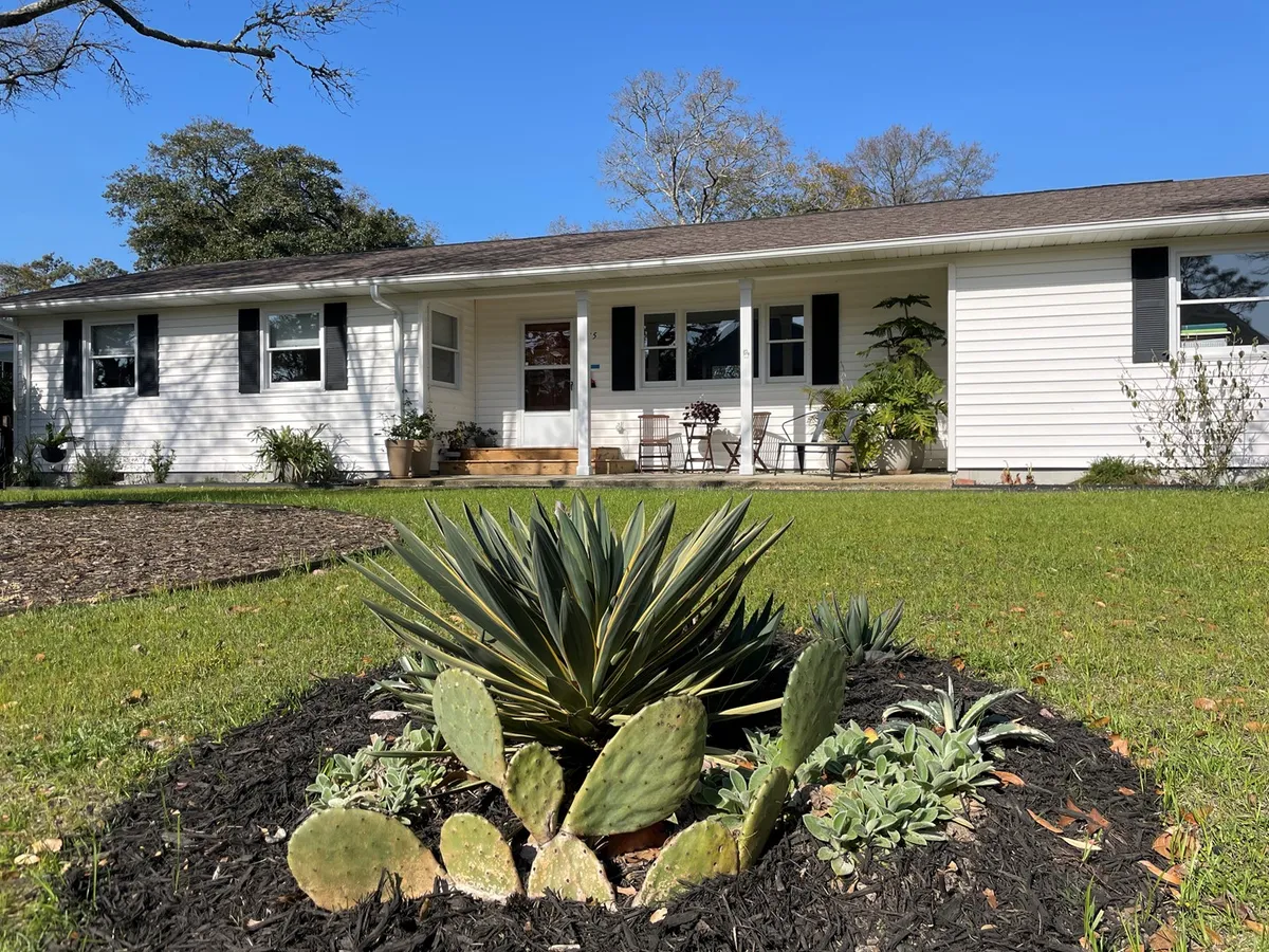 This exterior image features the inviting front porch of the house, bordered by a cactus garden and lush green grass. The porch offers a welcoming spot with seating, perfect for enjoying morning coffee while basking in the coastal breeze.