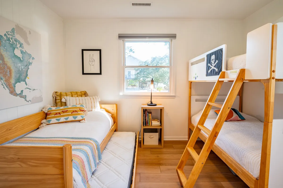 A playful and inviting bunk room featuring a twin-over-twin bunk bed, a single bed, a desk lamp, cheerful wall art, and a window letting in natural light. The arrangement is ideal for children, evoking both fun and comfort.