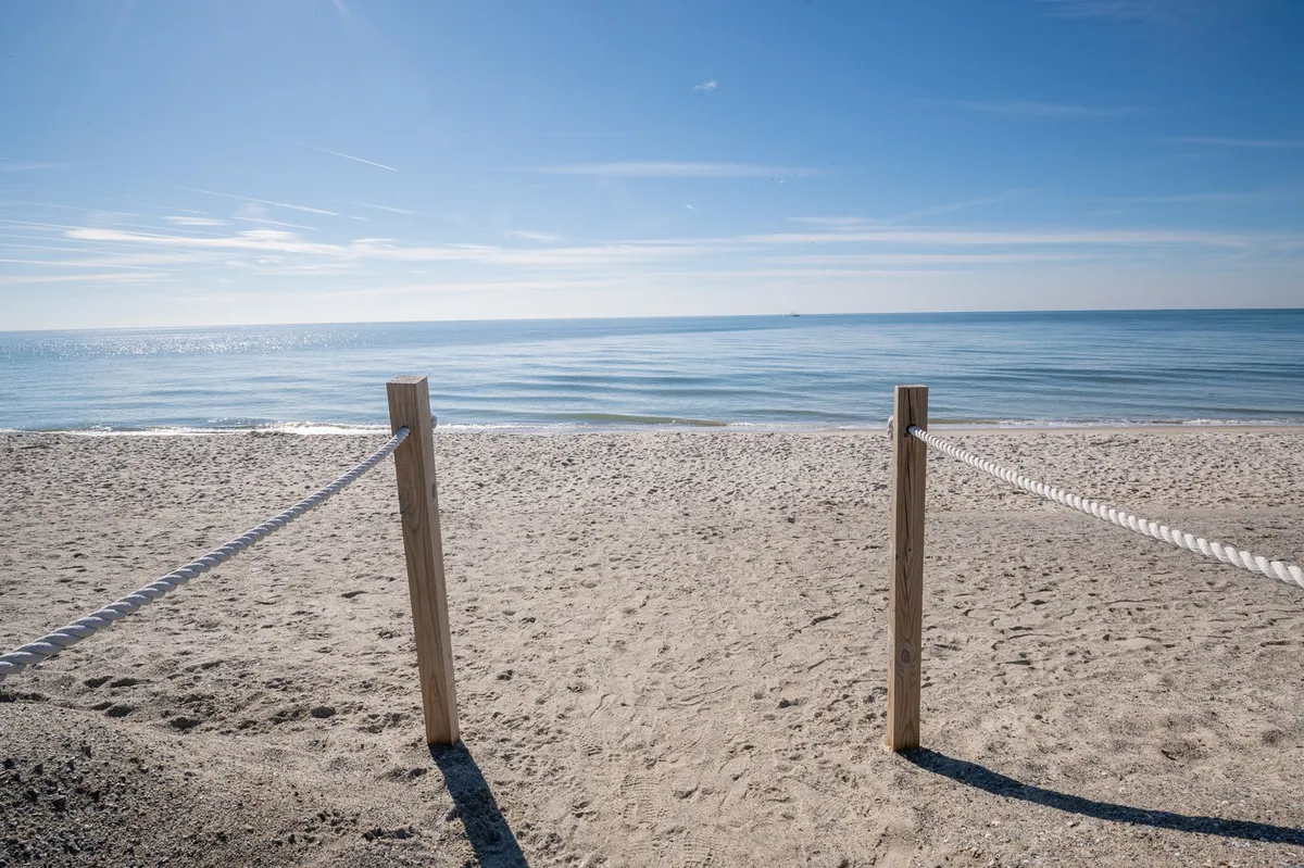 A serene view of a sandy beach bordered by wooden posts and a rope barrier, with the calm sea stretching into the distance. The scene suggests peaceful beach walks, sunbathing, and unwinding by the water—core activities for a relaxing short retreat.