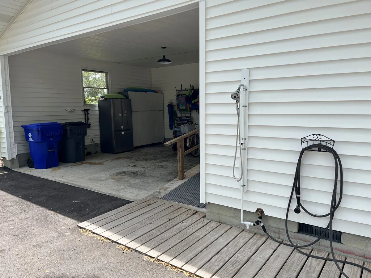 A tidy storage and mudroom area featuring blue trash bins, a gray storage cabinet, a white outdoor shower fixture, and wooden steps leading inside. The space is designed for convenient post-beach clean-up and easy access to gear and essentials.
